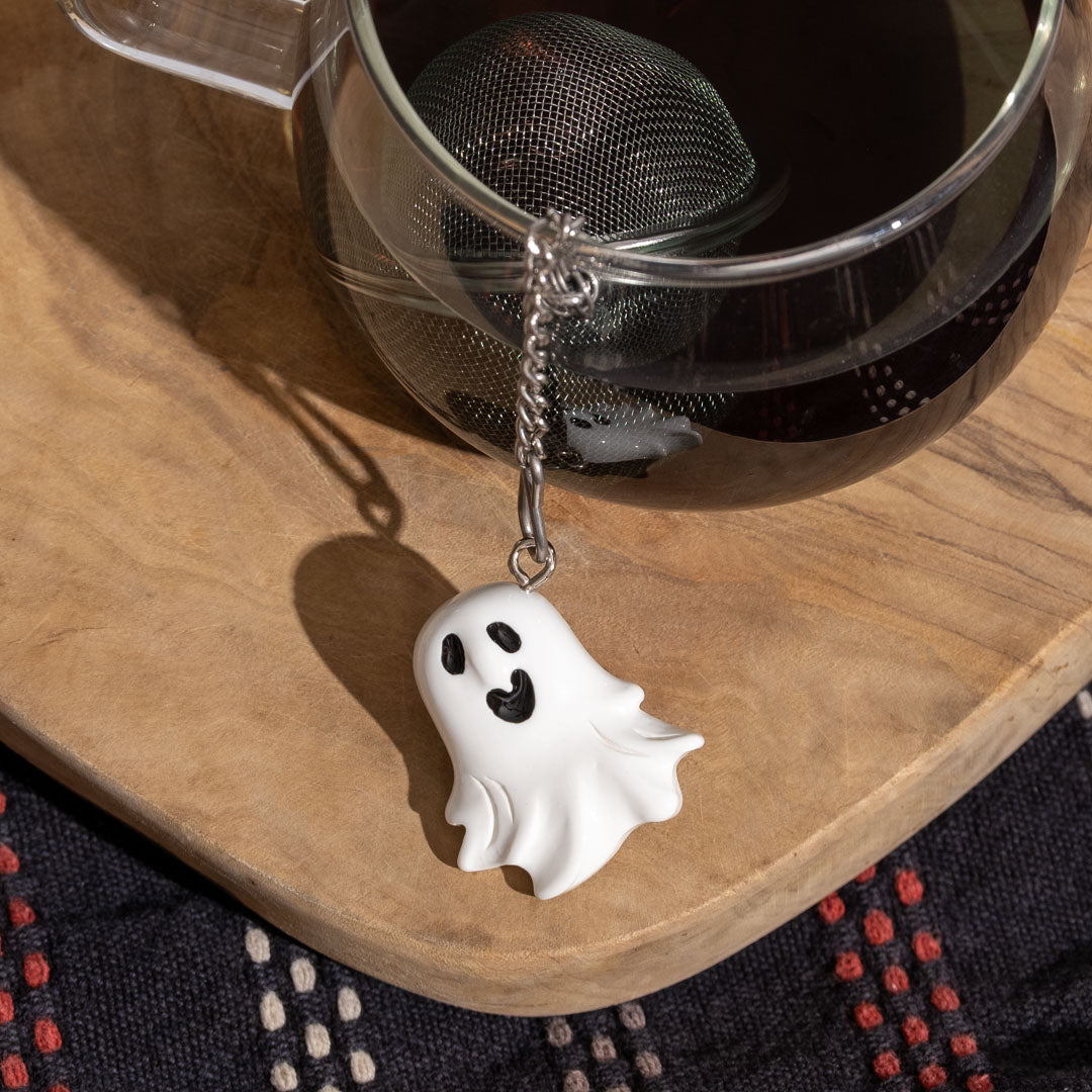 Ghost-shaped tea bag holder hanging from a teapot on a wooden surface.